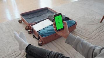 ung man Sammanträde på de golv i en mysigt levande rum och ser på de smartphone med en attrapp grön skärm stänga upp med en resväska med kläder som en bakgrund video
