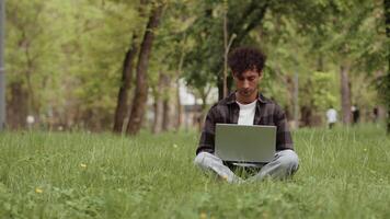 joven hombre en casual ropa trabajando con un ordenador portátil en un ciudad parque sentado en un césped, y luego aumento su manos y alegría debido a el éxito en su proyecto. concepto de flexible remoto trabajo video