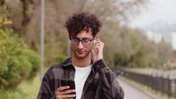 Young man in casual clothes and glasses close up walking in the city park with a smartphone, switching the music on his headphones and taking a rest after a work video