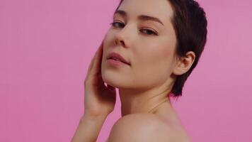 Young attractive woman with clean skin touching her face and coquettishly looking at the camera on a pink background close up. Concept of skin care for health and beauty video