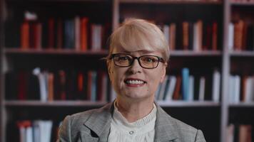 portret van een volwassen Kaukasisch vrouw in bril, staan, op zoek Bij de camera, en glimlachen met een backdrop van boekenkasten met veel boeken video