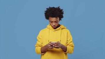 joven negro chico en pie y mecanografía un teléfono número en un teléfono inteligente y empieza a hablar con un amigo con un sonriente cara en un azul antecedentes en el estudio video