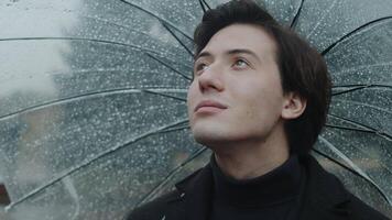 Young man in a coat standing under a wet transparent umbrella in the city park close up, looking up at cloudy skies and feeling moments of autumn video