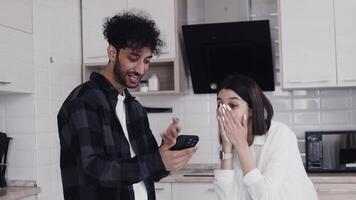 Young woman looking at the smartphone of her partner, becoming surprised and overjoyed, and they are starting to hug together in cozy kitchen at home video