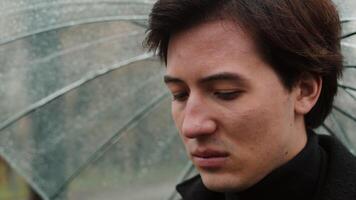 Young man in a coat standing under a transparent umbrella close up and blowing his nose into a handkerchief in the autumn city park in rainy weather video