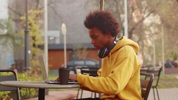 jovem Preto programador bebendo café a partir de uma papel copo e trabalhando em uma computador portátil, sentado às a mesa dentro a ao ar livre cafeteria em uma cidade rua em uma ensolarado dia video