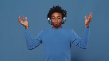 jong zwart vent in hoofdtelefoons proberen naar kom tot rust en beginnend naar mediteren Aan een blauw achtergrond in een studio. Mens Verbinden de duim en wijsvinger Aan beide handen en zetten hen naar de zijden video