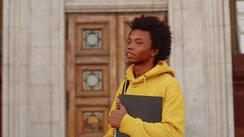 Young black programmer in a casual clothes with a laptop and backpack looking into the distance, then smiling at the camera with a building as a backdrop video