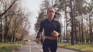 Young caucasian female athlete in a black sportswear running cheerfully on an alley in an autumn city park on a sunny day, front view. Concept of modern outdoor sports training in a casual urban life video