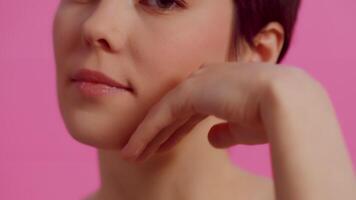 Young attractive woman with clean skin touching her face from chin to cheek and gently looking at the camera on a pink background close up. Concept of skin care for health and beauty video