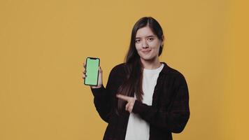 joven atractivo mujer demostración un teléfono inteligente con un Bosquejo verde pantalla y alegremente señalando a eso con un dedo en un amarillo antecedentes en el estudio. concepto de Consejo y promoción video