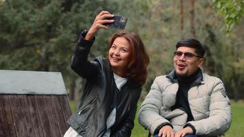 deaktiviert asiatisch Mann im ein Rollstuhl und asiatisch Frau lächelnd und haben Spaß zusammen während nehmen ein Joint Selfie auf ein Smartphone im ein Herbst Stadt Park video