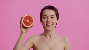 Young attractive woman with clean skin, covering her own eye with a half of the grapefruit and smiling on the pink background. Concept of skin care for health and beauty video