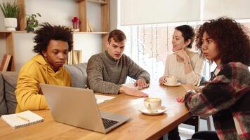 Young corporate employee presenting his project to multiracial colleagues and pointing with a finger to a screen of a laptop while sitting together in office on the common table with laptop video