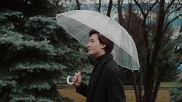 Young man in a coat unhurriedly walking under a transparent umbrella in the autumn city park, looking around and smiling despite the rainy weather video