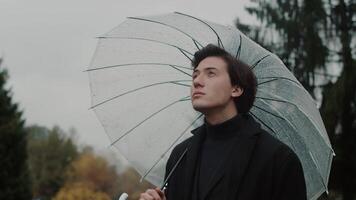 Young man in a coat standing under a transparent umbrella in the autumn city park, looking up at cloudy skies and smiling despite the rainy weather video