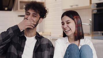 Young cheerful couple in casual clothes sitting on a couch in a kitchen and laughing loud when looking at the camera. Concept of happiness and joy in the relationship video