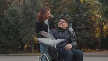 Disabled asian man in a wheelchair feeding pigeons in an autumn city park and laughing together with the asian woman. Concept of acceptance, love and support for people with disabilities video