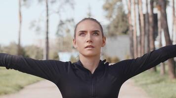 Young attractive caucasian sportswoman raises her hands up, performing warming up exercise before a run and tuning up for a good workout on an alley in an autumn city park close up video