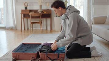 Young man neatly folding his clothes into a suitcase lying on the floor at home and zipping it during attentive preparation for the vacation video