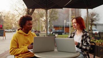 ung caucasian flicka förklara en ung svart kille handla om hans studie frågor. ung människor i tillfällig kläder Sammanträde med bärbara datorer i de utomhus- Kafé, talande och dricka kaffe video
