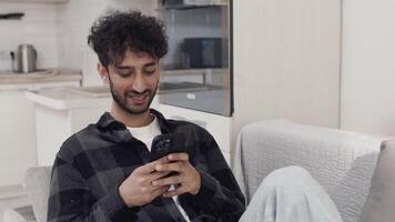 Young man sitting on a couch in a cozy kitchen at home and scrolling the feed on his smartphone fast. Guy consuming a large amount of content video