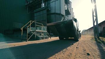 Loader Moving at Industrial Facility, A large loader is moving through an industrial facility with silos and various structures visible, creating a dusty atmosphere as it drives. video