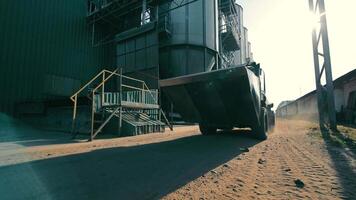 Lader ziehen um beim industriell Einrichtung, ein groß Lader ist ziehen um durch ein industriell Einrichtung mit Silos und verschiedene Strukturen sichtbar, Erstellen ein staubig Atmosphäre wie es fährt. video