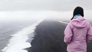 enda kvinna turist stå omtänksam se på atlanten hav vågor. känd ikoniska klippa synpunkt över reynisfjara svart sand strand. person utseende för riktning och ändamål på resor video