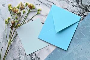 A blue envelope and a blank notecard sit on a marble table next to a sprig of yellow flowers. photo