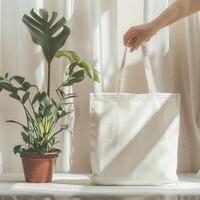 A simple tote bag made of beige canvas. The bag is empty and a hand is holding the handles. There is a potted plant next to the bag. photo