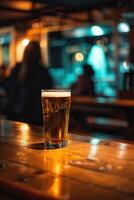 A half-full glass of beer sits on a wooden bar. There are people in the background. photo