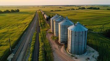 A railroad track runs through a rural area, passing by a group of grain silos. photo