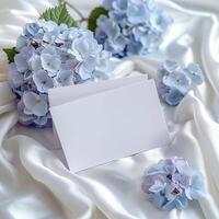 azul y blanco tema. un blanco Nota tarjeta se sienta en un cama de suave blanco y azul seda. azul flores con un blanco antecedentes. Perfecto para un Boda o especial ocasión. foto