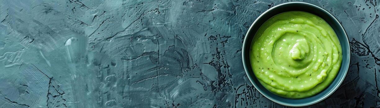 A close-up top view of a bowl of creamy avocado soup, vibrant green color in focus, set against a muted, contemporary background, providing a wide, panoramic space for text. photo