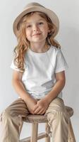 blank unisex Canvas toddler T-shirt Mockup, color white, girl model sitting on a stool wearing khaki pants and a hat photo