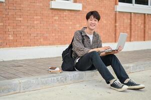 un contento asiático masculino Universidad estudiante se sienta en el calle cerca el instalaciones edificio utilizando su computadora. foto