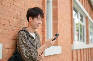 un contento asiático masculino Universidad estudiante soportes en contra un ladrillo pared de un edificio utilizando su teléfono inteligente foto