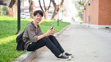 un amigable, positivo joven asiático masculino Universidad estudiante se sienta en el calle por el instalaciones edificio. foto