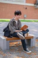 un feliz, relajado asiático masculino Universidad estudiante se sienta en un banco en el instalaciones parque leyendo un libro de texto. foto