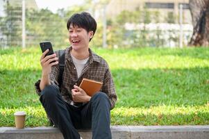 A cheerful Asian male college student sits on the street using his phone, laughing at messages. photo