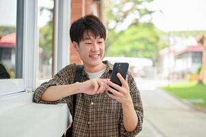 un sonriente joven asiático masculino Universidad estudiante soportes por el instalaciones edificio utilizando su teléfono inteligente foto