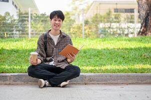 Smiling Asian student sits on the street in the campus park with a coffee cup and a book in his hand photo