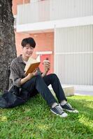 un feliz, relajado asiático masculino Universidad estudiante disfruta leyendo un libro debajo el árbol en el instalaciones parque. foto