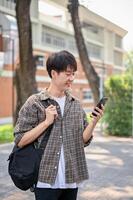 A happy Asian male college student is using his smartphone while walking in the campus park. photo