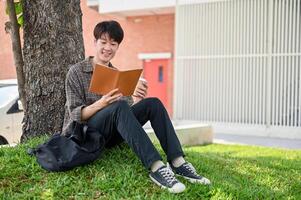 un feliz, relajado asiático masculino Universidad estudiante disfruta leyendo un libro debajo el árbol en el instalaciones parque. foto