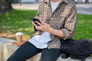 A cropped image of a male college student sits on a stone bench in a park, using his smartphone. photo