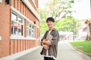 A cheerful Asian man student stands outside a brick building on campus, holding a laptop and books. photo