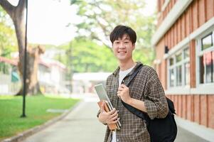 A cheerful Asian man student stands outside a brick building on campus, holding a laptop and books. photo