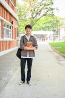 A positive Asian man college student, stands in front of a brick building on campus, holding books. photo